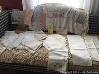 Lot of vintage table linens including napkins, fingertip towels, table runners and tablecloths arranged on and in front of a couch with plaid upholstery.
