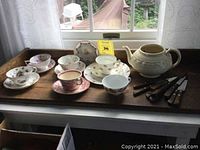 Overall view of 6 floral cups and saucers, 2 extra plates, Adams Royal ivory Titian Ware teapot, and cutlery on a wooden tray by window.