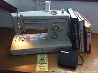 Front and side view of Kenmore sewing machine with labeled model number, lit needle area, spool of white thread on top and foot pedal beside it, with five DIY books upright behind it.