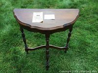View of the hardwood demilune table from front to show scalloped half-moon surface and three turned legs connected by a base stretcher.