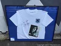 Framed white Gloria Estefan autographed T-shirt with photo in frame showing full display and cracked glass.