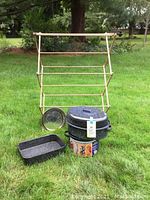 Photo showing wooden vintage foldable drying rack on grass with blue and white speckled enamelware cookware items including roasting pan, pot with lid, and rectangular dish, with a can used for display underneath pot.