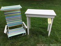 Outdoor photo showing white painted wooden table and striped folding chair with canopy on grass.