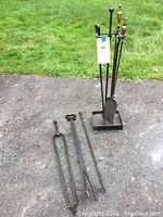 Photo shows entire fireplace tool set including stand and 4 tools (tongs, 2 pokers, shovel) on gravel next to grass.