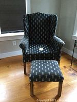 Full view of traditional wing chair with matching hassock, pineapple patterned dark upholstery with gold motifs, solid wood legs.