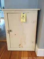 Front view of rustic beige/ivory cupboard with a single door and knob, showing chipping paint and wear on door.