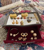 Mirrored jewelry box with red velvet drawer opened showing various earrings and brooches arranged inside and on top.