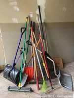 Image showing assortment of snow shovels, leaf rakes, broom, and push leaf sweeper placed in garage corner against wall