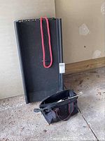 Black plastic foldable dog ramp with red carrying strap, standing upright against wall, tool bag on floor in front.