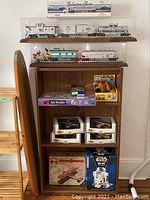Front view of wooden bookcase showing all contents: model trains in display cases on upper shelves, boxed model planes on middle shelf, plush bear and boxed games (Yahtzee, Art Studio) below, Star Wars R2D2 phone at bottom.