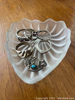 Five sterling silver rings and Kokopelli brooch displayed inside frosted glass heart-shaped trinket box.