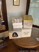 Loose and boxed Department 56 Snow Village figurines stacked on a round table, some in original packaging and styrofoam inserts.