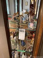 Front view of glass cabinet displaying various Norman Rockwell figurines including children with animals, individuals seated at desks, and a child riding a horse.