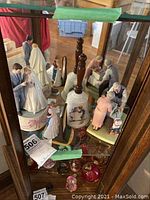 Collection of Norman Rockwell wedding themed figurines, music box, and Hummel bell displayed in cabinet with reflections