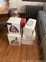 Original boxes for multiple Department 56 Snow Village buildings and Lionel Electric Trains stacked on wooden floor.