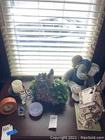 Photo of items on dark wood table near window with blinds. Items include a small clock, candle holder with candle, Blue Sky church candle holder, glass bowl, two small vases, lighthouse figurine, plush elephant, and a house figurine.