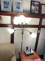 Front view of multi-head floor lamp with all five shades illuminated, positioned behind a chair and adjacent to a wooden side table with phones and other items.