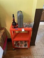 View of orange metal rolling cart with Skil cordless screwdriver case, plastic bin with tools and screwdrivers on top shelf.