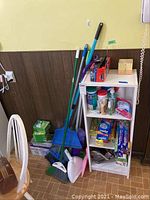 Wide photo showing the shelving unit with cleaning supplies and multiple brooms and dust pans leaning against it.
