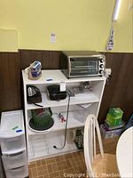 Full view of white shelving unit with Black & Decker toaster oven on top, Sony clock radio and another radio on middle shelf, plastic green basket on bottom shelf. Batteries and battery organizer on middle shelf. White plastic drawers and green box beside bookshelf.