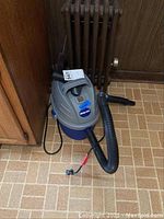 Shop Vac wet/dry vacuum on a tiled floor near wooden cabinets and a radiator, showing front and side view with hose and power cord.