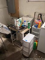 Showing metal shelving unit with tiled surface, plastic drawers nearby, and various cleaning products arranged on and around it in a basement laundry area.