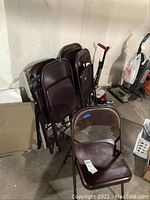 Eight brown metal folding chairs stacked and one chair set up, located in a basement storage area with concrete floor and walls.