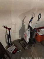 Photo showing floor polisher standing next to two vacuum cleaners in basement corner.