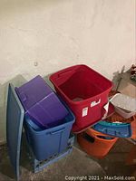 View of assorted colored Sterilite storage totes with some lids off and some lids beside the containers in basement setting.