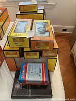Stack of Rail King and MTH original boxed electric train sets on bed, showing orange and yellow boxes with Rail King branding and some box window displays