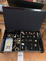 Open black jewelry box showing multiple compartments with jewelry items including brooches, earrings, rings and a religious image card