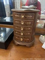 Front view of oval wooden jewelry box showing five drawers with floral and gold designs, round gold knobs, sitting on a vanity tabletop.
