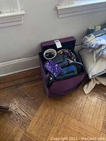 Purple lidded storage ottoman open, displaying contents including handbags, eyeglasses, sunglasses, and a planner.