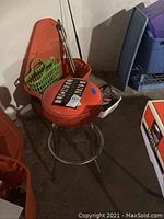 Front and side view of the orange Lionel stool with round padded seat and chrome metal frame with four legs and circular footrest, placed on basement floor near orange storage bins.