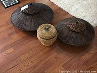 Three wicker baskets with lids on wooden floor, two dark brown large round baskets and one smaller natural tan basket.