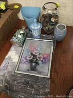 Photo showing two rugs beneath a framed floral painting, three vases in blue, clear and ceramic styles, and a box of decorative shells and stones.