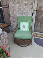Front view of metal wicker swivel chair with green cushions and floral pillow, showing the chair on a front porch area with brick wall background.