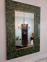 Large rectangular mirror with beveled edges and green-blue tile mosaic frame, installed on wall, reflecting dining room chandelier and table
