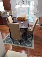 Dining table with four beige upholstered chairs around it, sitting on a blue patterned rug in a kitchen area.