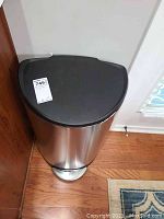 Front angled view of chrome trash can showing black lid with lot sticker and foot pedal on wooden floor