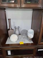 Display of various decorative vases, a white textured orb, a spherical mosaic object, a lead crystal dog figurine, and a modern glass container inside a wooden cabinet.