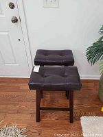 Pair of brown leather padded stools with tufted seats and dark wooden legs shown side by side against a white wall and hardwood floor.