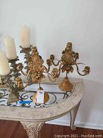 Group shot showing brass floral candelabra and multiple ornate candle holders with pillar candles on glass-top decorative table.