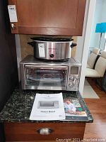 Emeril Lagasse stainless steel air fryer toaster oven with Hamilton Beach crock pot on top, with accompanying manual and cookbook.