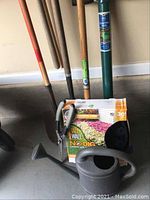 Photo showing garden clippers with grey handles, planting tool with green handle grip, and box of landscape edging with spikes in original packaging, alongside long-handled tools.