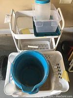White metal rolling cart with three mesh shelves holding a clear plastic bin and a large blue bucket, a white laundry basket with a blue bucket inside it is in front of the cart.