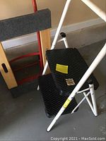 Side view of white two-step stepladder with black steps and red hand truck partially visible