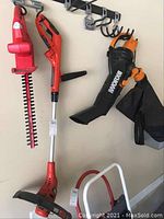 Three electric yard tools hung on garage rack: Black & Decker trimmer, Homelite hedge trimmer, WORX leaf blower with collection bag.