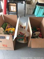 Two open cardboard boxes containing various lawn and garden chemical bags and bottles, mostly partially used.