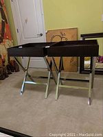 Two metal folding tray frames side by side on carpet, brown leather trays with crocodile pattern on top, shiny polished metal legs crossed in X shape.